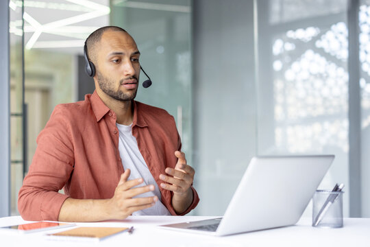 A man in a headset is looking at a laptop and gesturing with his hands during a video call in a modern office setting.