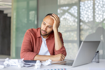 A frustrated man with a hand on his head, surrounded by crumpled papers and a laptop. He looks tired and stressed.