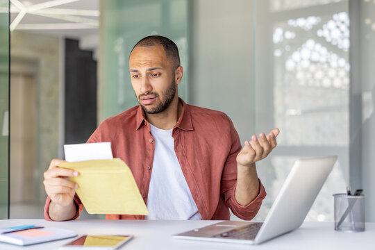 A man appears confused while looking at a letter, seated at a desk with a laptop in front of him.