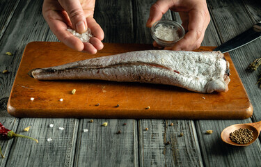 A person seasons a fresh fish fillet with salt while holding a small jar of spices, set on a rustic...