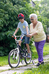 Obraz premium Grandmother Guides Grandson Learning to Ride Bicycle with Training Wheels. Family Bonding, Child Development, Active Summer Outdoor Activity. Copyspace