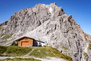 Wooden hut at the base of a steep gray cliff in the Alps.