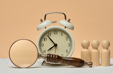 White vintage alarm clock, magnifying glass and three wooden human figures. The composition symbolizes the search for time, efficiency or analysis of resources.