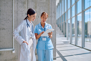 Female doctor and nurse reviewing patient information on digital tablet in bright hospital corridor