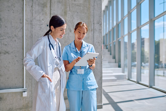 Female doctor and nurse reviewing patient information on digital tablet in bright hospital corridor