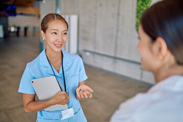 Two young nurses are standing in a bright, clean hospital corridor, engaging in conversation while holding a tablet to share information
