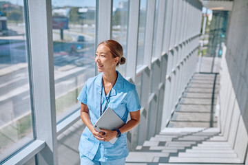 Smiling young female doctor in blue uniform walking up stairs in modern hospital holding tablet