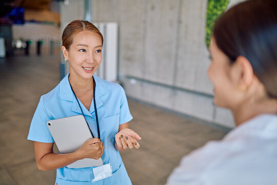 Two young nurses are standing in a bright, clean hospital corridor, engaging in conversation while holding a tablet to share information