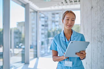 Smiling healthcare worker in blue uniform using a digital tablet in a bright, modern clinic hallway, showcasing technology in healthcare