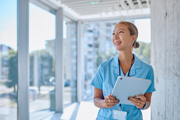 Smiling asian female doctor wearing blue uniform holding tablet and looking away in hospital...