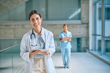 Portrait of a smiling female doctor using a tablet in a hospital corridor, with a nurse walking in the background