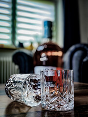 Two empty crystal glasses on a wooden table in front of a sofa and a bottle, with blurred window blinds in the background.