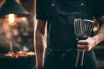 Man in Black Chef Apron Holding Barbecue Tongs and Spatula