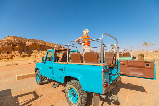 Blonde tourist standing at Jabal Al-Ahmar on a vintage 4x4 car, admiring the stunning view of the Hegra archaeological site in Al-Ula, Saudi Arabia, under a clear blue sky