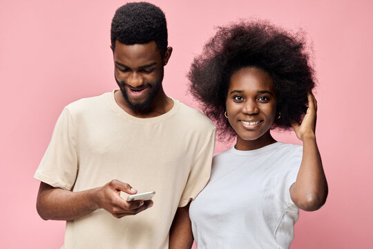 Happy young couple with Afro hairstyles, smiling and looking at a smartphone against a pink background, representing love and technology connection