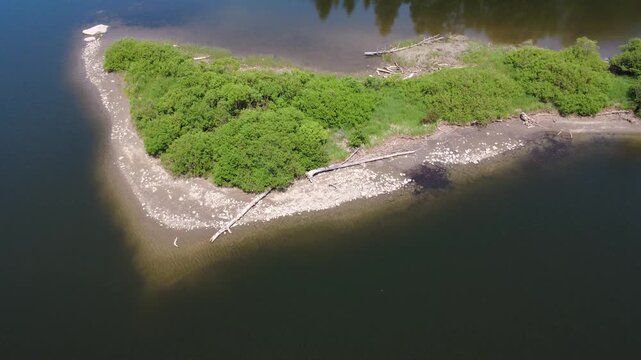 Serene landscape along the Mitis River with a lush island covered in shrubs and driftwood, peaceful water reflections, and the Mitis-2 hydroelectric dam visible in the background. Quebec, Canada.