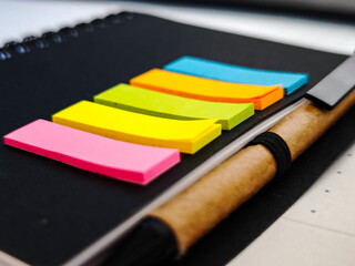 Notebook with colorful sticky flags next to a wooden pen. All placed on a dark surface.