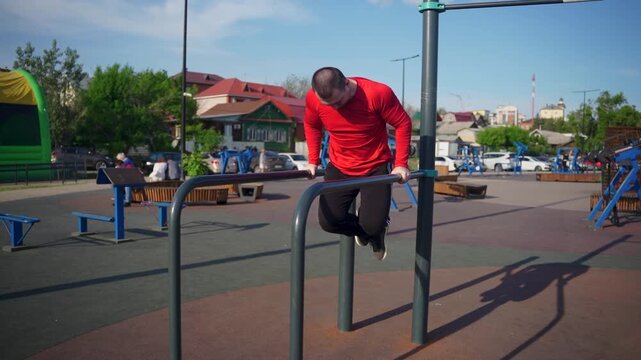 Man in red shirt doing dips on parallel bars at outdoor gym. He looks down.