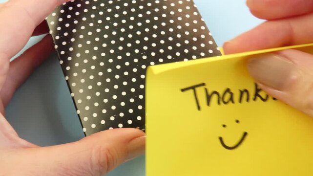 Woman hands add a yellow sticky note to a polka dot gift box. Sticky note with the message "Thanks" and a smile.