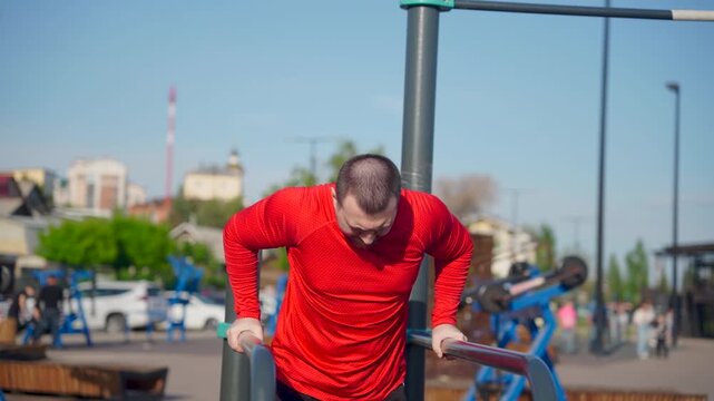 Man in red shirt doing dips on parallel bars at outdoor gym. He looks down.