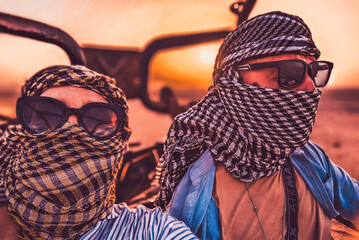 Two people with traditional scarves and sunglasses sit ready in an off-road vehicle.