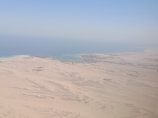 High-altitude photograph showing a vast desert landscape meeting the clear blue waters of the sea. Taken from an airplane window, the image captures sandy textures