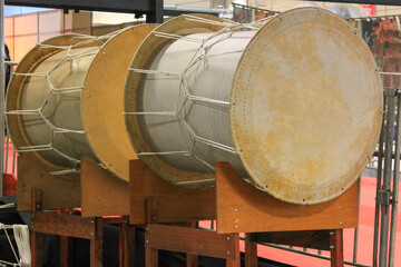 Two large Taiko drums, with two light skins and wooden sides, are arranged on wooden stands. They...