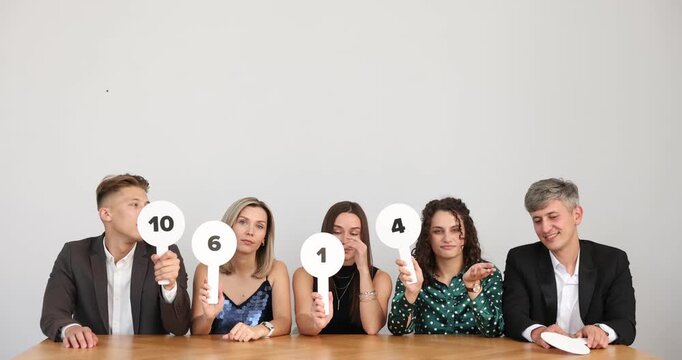 Panel of emotional judges voting with score signs at table near grey wall