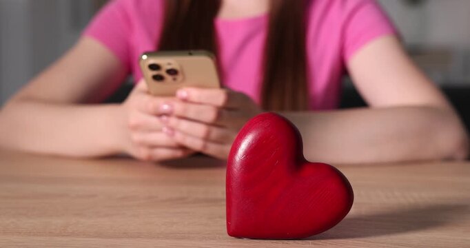 Online dating site. Woman using smartphone at wooden table indoors, focus on decorative heart