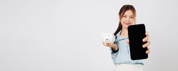 Smiling Asian woman holding mobile phone and white piggy bank with joy showing bank application...