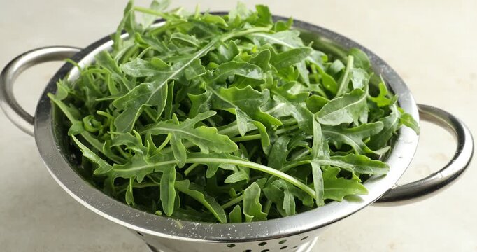 Spraying water onto fresh ripe green arugula leaves in metal colander on white table, closeup