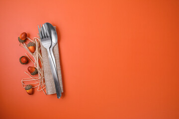 Autumn flat lay composition with stainless steel fork and spoon wrapped in a burlap napkin, surrounded by decorative acorns and straw on a vibrant orange background. 