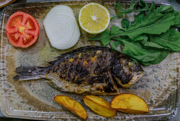Grilled whole sea bream, garnished with tomato, onion, lemon, arugula and potatoes, served on a textured platter at a restaurant in Istanbul, Turkey