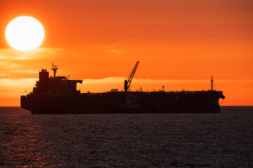 Silhouette of a large cargo ship, oil tanker sailing on the ocean, dramatic orange sunset, sun over...