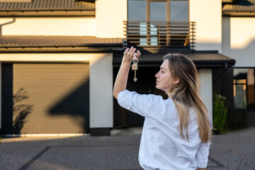 Happy woman standing with house keys in front of residential home. Real estate, moving home or...