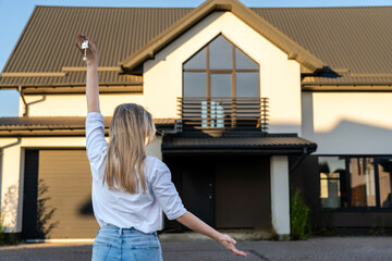 Happy woman standing with house keys in front of residential home. Real estate, moving home or renting property concept
