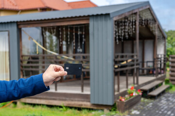 Plastic credit card in hand with new house at background. Broker, mortgage, property, real estate concept. Buying or renting