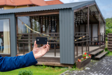 Plastic credit card in hand with new house at background. Broker, mortgage, property, real estate concept. Buying or renting