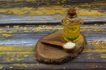 Glass bottle of sesame oil and raw sesame seeds in wooden spoon or bowl on rustic table. Uncooked...