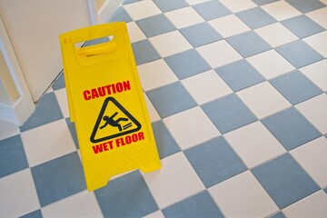 Yellow caution wet floor sign standing on a checkered blue and white tiled floor, warning of...