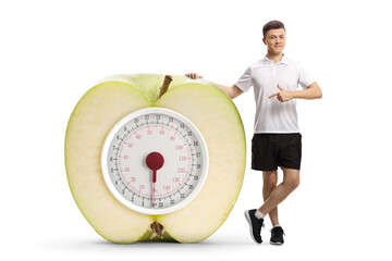 Full length portrait of a young man pointing at a big apple with a weight scale