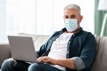 Senior grey-haired man in protective face mask sitting on sofa in living room, using modern laptop,...