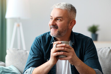 Relaxed senior grey-haired man with mug of coffee or tea sitting on sofa at home, looking at copy space and smiling. Joyful matured man drinking tea in living room. Leisure, domestic lifestyle concept