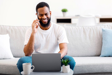 Always In Touch. African Guy Working On Laptop And Having Phone Conversation Doing Job Remotely...