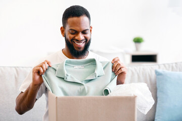 Shopping Delivery. Joyful African American Man Receiving Clothes Unpacking Cardboard Box And...