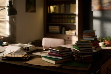 Stack of assorted books arranged on desk in home office with sunlight streaming through window,...