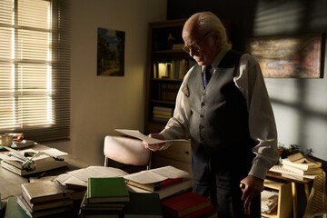 Senior Caucasian man with hair and mustache standing by desk reading document, wearing glasses and...