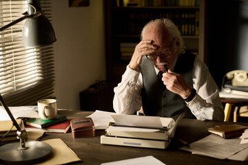 Senior Caucasian man sitting at desk reading document while holding glasses and pen, appearing focused and thoughtful, surrounded by paperwork and vintage typewriter in office setting
