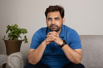 Indian young man looking at camera while sitting on cozy sofa at home. wearing blue casual t-shirt...