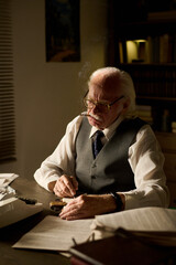 Senior Caucasian man with hair and mustache sitting at desk reviewing documents and holding cigar,...